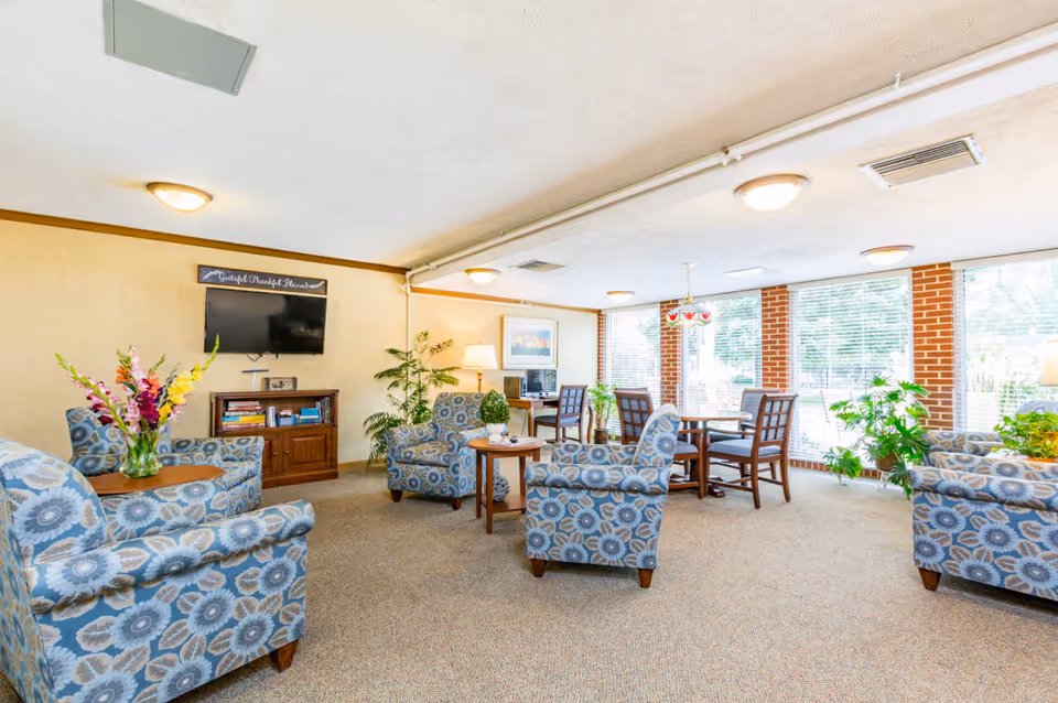 Bright communal lounge with patterned armchairs, small tables, a wall-mounted TV, and a dining table by large windows.