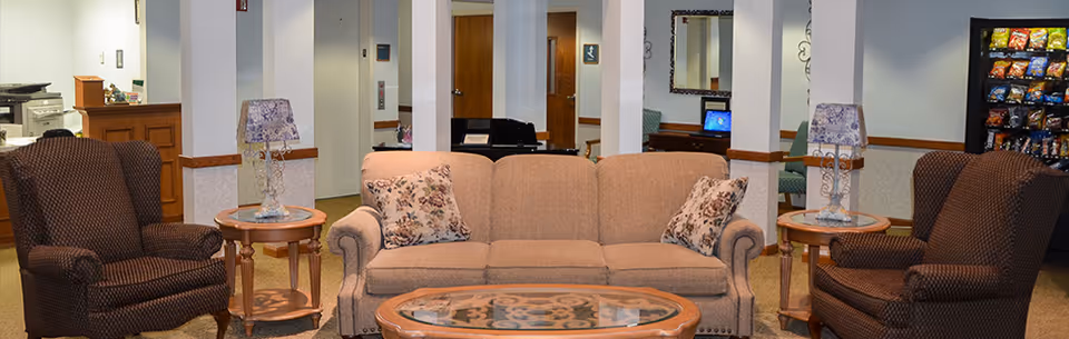 A cozy living room area in a senior living facility featuring a beige sofa with floral cushions, two brown armchairs, two side tables with decorative lamps, a glass coffee table with a patterned design, and a vending machine in the background.