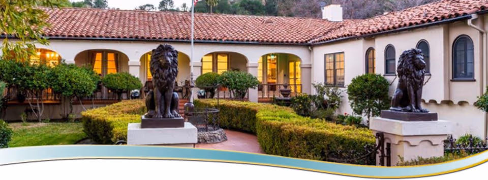 Outdoor courtyard area of a retirement facility with trimmed hedges, two large lion statues on pedestals, and a building with arched windows and a red tile roof in the background during dusk.
