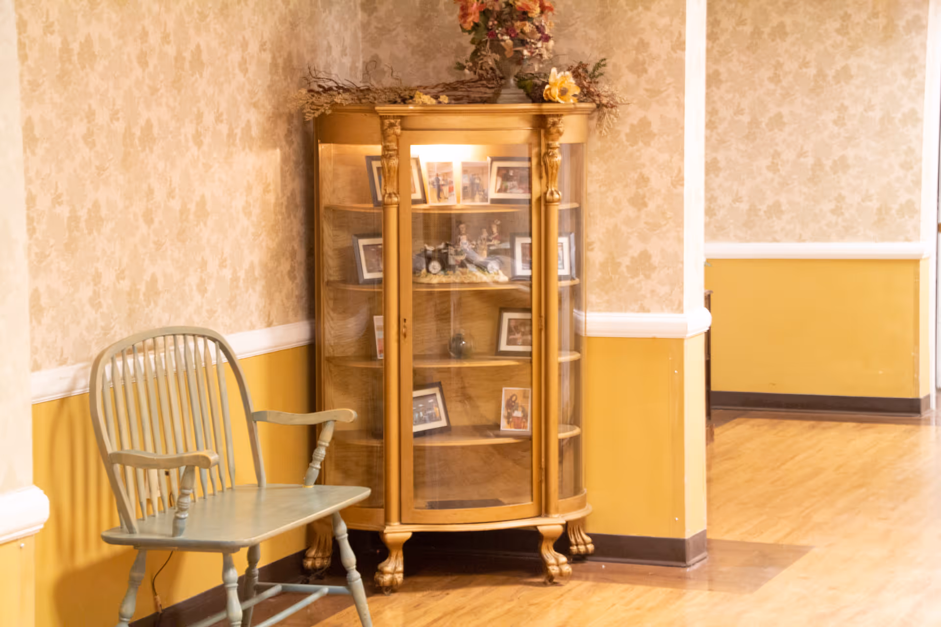 A corner of an interior hallway with yellow and beige floral wallpaper and wood flooring. There is a light green wooden bench on the left and a wooden display cabinet with glass doors containing framed photos and decorative items. A floral arrangement is on top of the cabinet.
