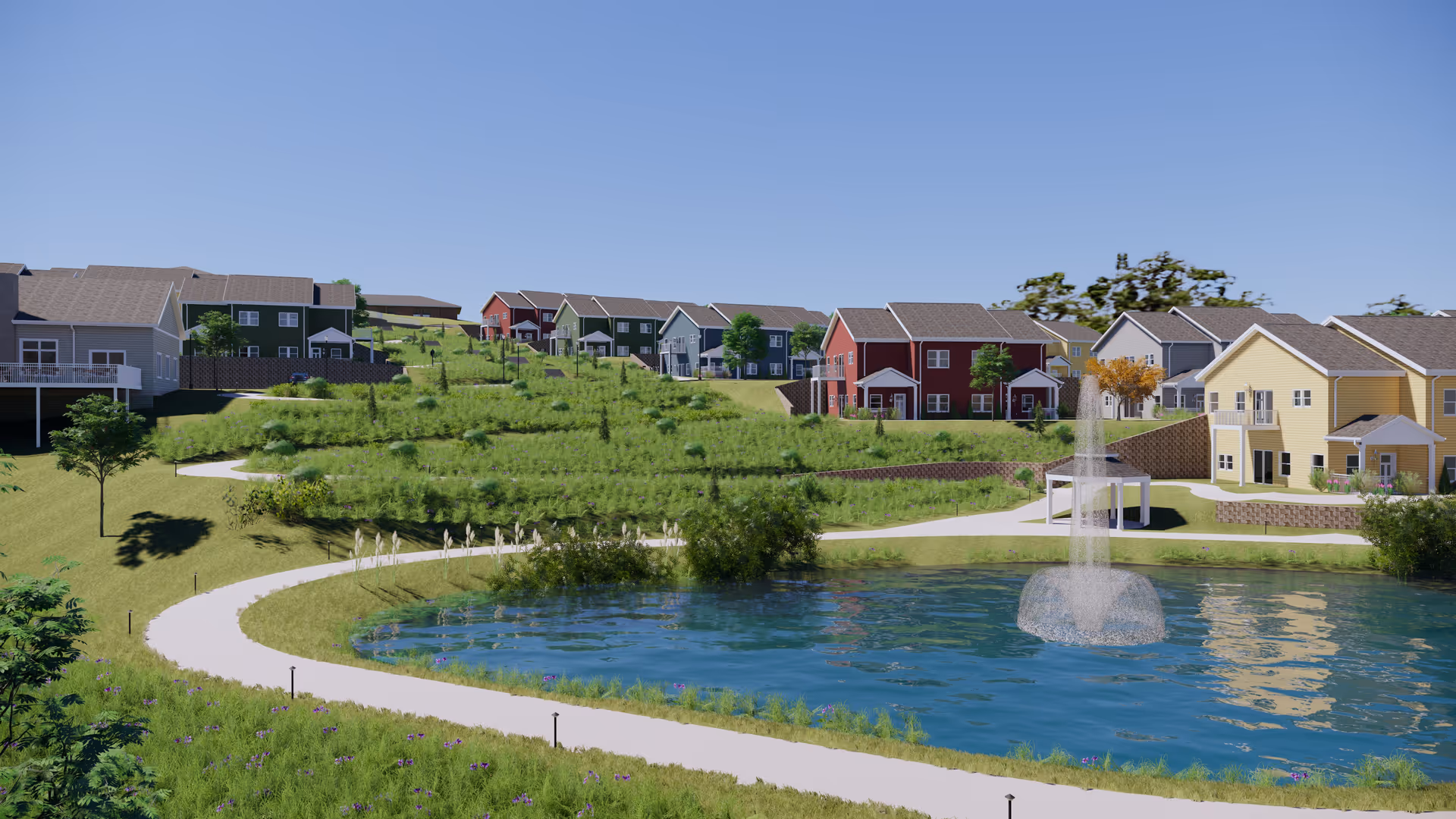 A scenic outdoor view of a senior living community with colorful houses on a grassy hillside surrounding a pond with a water fountain. A curved walking path winds around the pond, and there are trees and shrubs scattered throughout the landscape under a clear blue sky.
