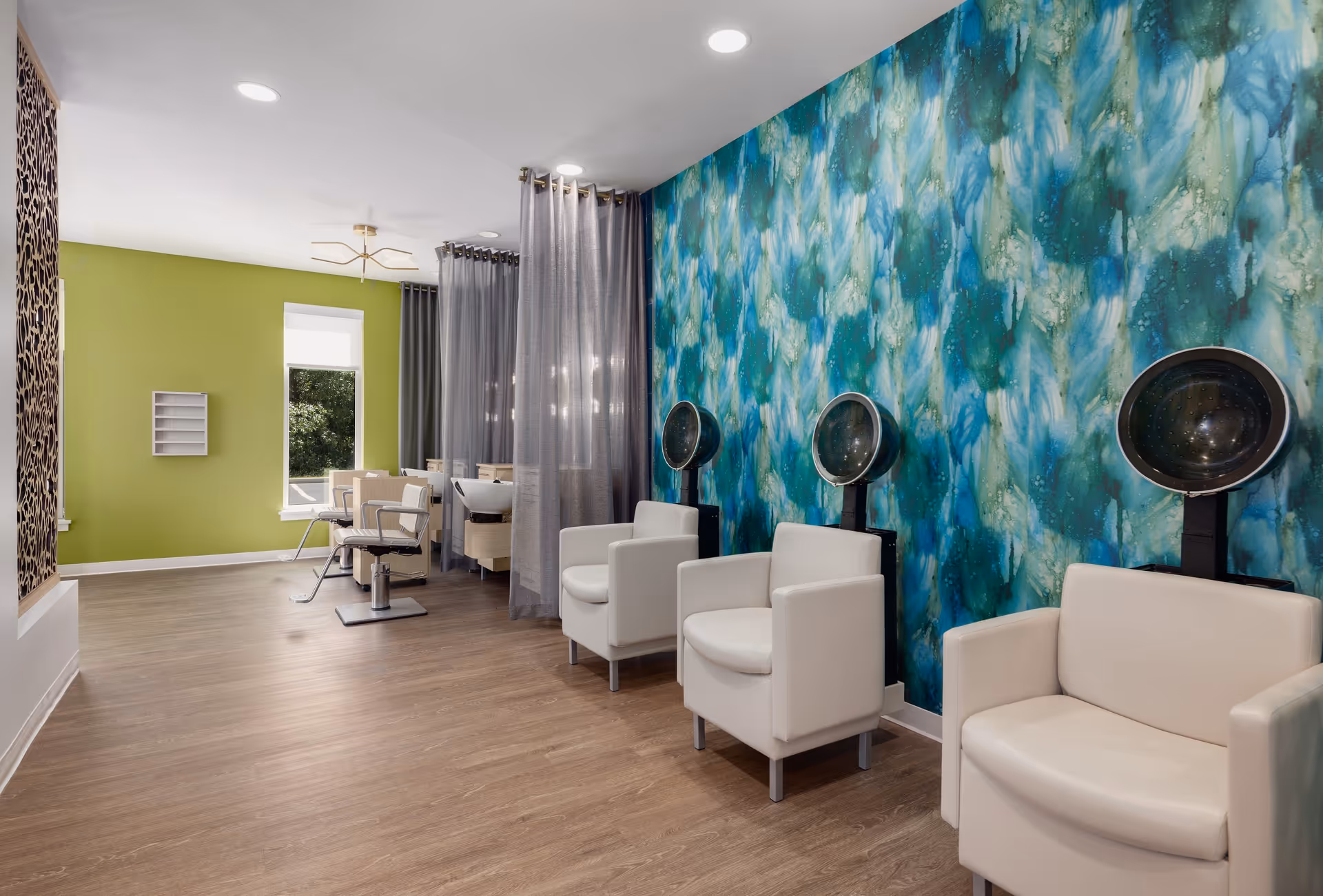Interior of a salon area in a senior living facility with three white salon chairs under hair dryers against a blue and green abstract patterned wall. The room has wood flooring, a green accent wall with a window, and additional salon chairs separated by sheer gray curtains.