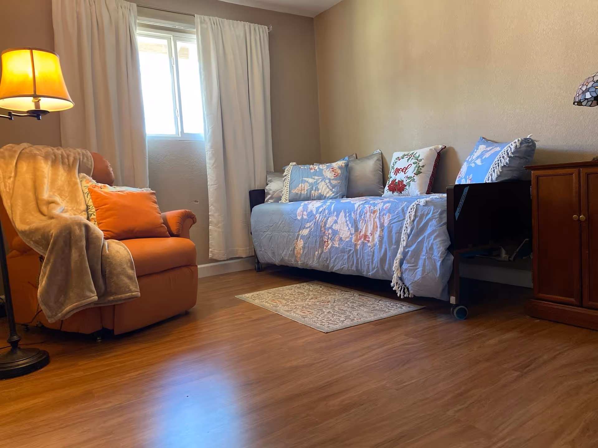 A cozy room with a wooden floor featuring a daybed covered with a light blue floral quilt and multiple pillows, including one with a 'Noel' design. To the left, there is an orange armchair draped with a beige blanket and an orange cushion. A floor lamp with a yellow shade stands beside the armchair. A small wooden cabinet with a stained glass lamp on top is on the right side of the daybed. A window with white curtains lets in natural light.
