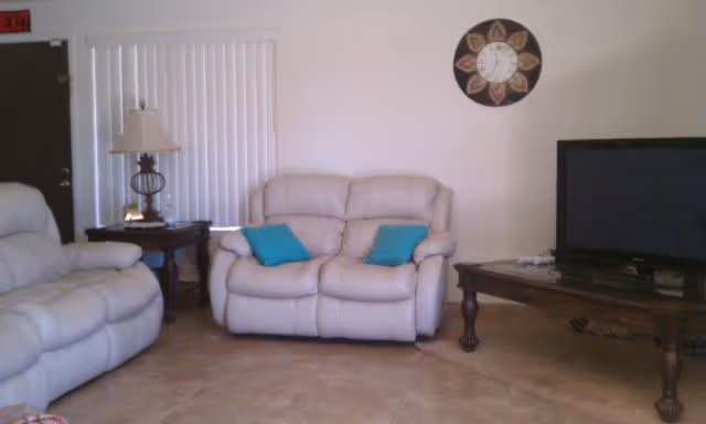 A cozy living room with two beige recliner sofas, each with blue throw pillows. There is a wooden side table with a lamp between the sofas, vertical blinds on the window behind, a decorative wall clock, and a wooden TV stand with a flat-screen television.