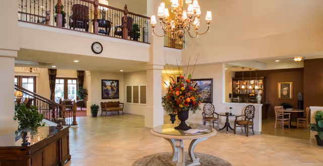 Spacious and well-lit senior living facility lobby with a round table holding a large floral arrangement in the center. There is a chandelier hanging from the ceiling, a staircase with wooden railings leading to an upper floor, and seating areas with chairs and tables. The walls are decorated with framed artwork and plants are placed around the room.