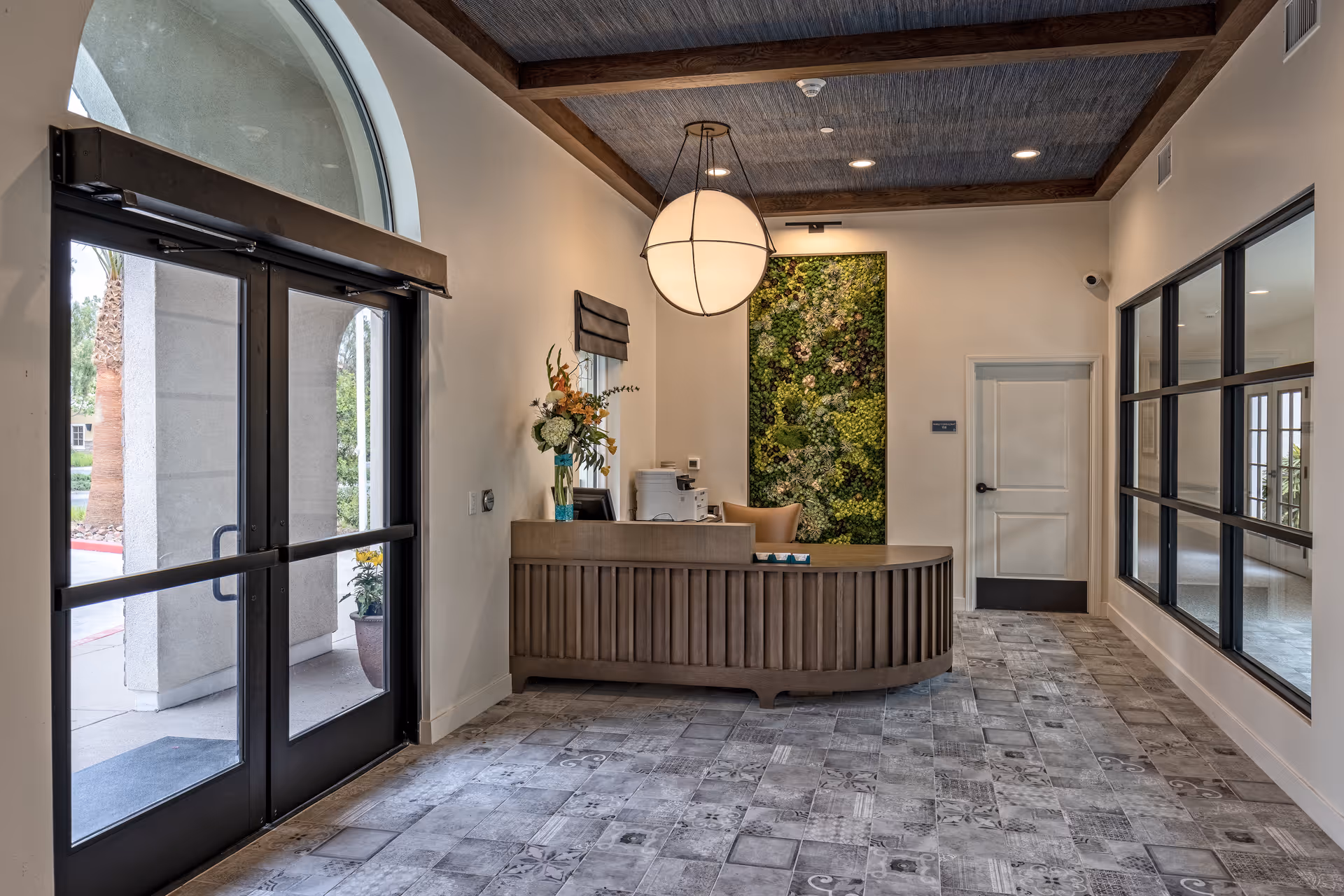 Reception area of Serra Sol Memory Care featuring a curved wooden front desk with a chair behind it, a large round hanging light fixture above, a vertical green plant wall behind the desk, a vase with flowers on the desk, large glass double doors on the left, and a window with black frames on the right.