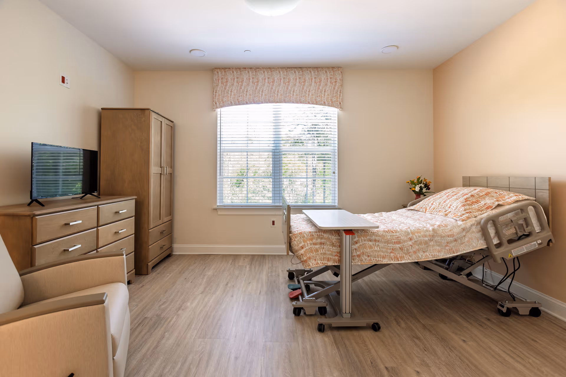 A bright and clean bedroom in a senior living facility with a hospital-style adjustable bed covered in a patterned bedspread, a small overbed table, a wooden dresser with a flat-screen TV on top, a wooden wardrobe, a beige armchair, and a large window with blinds and a valance letting in natural light.