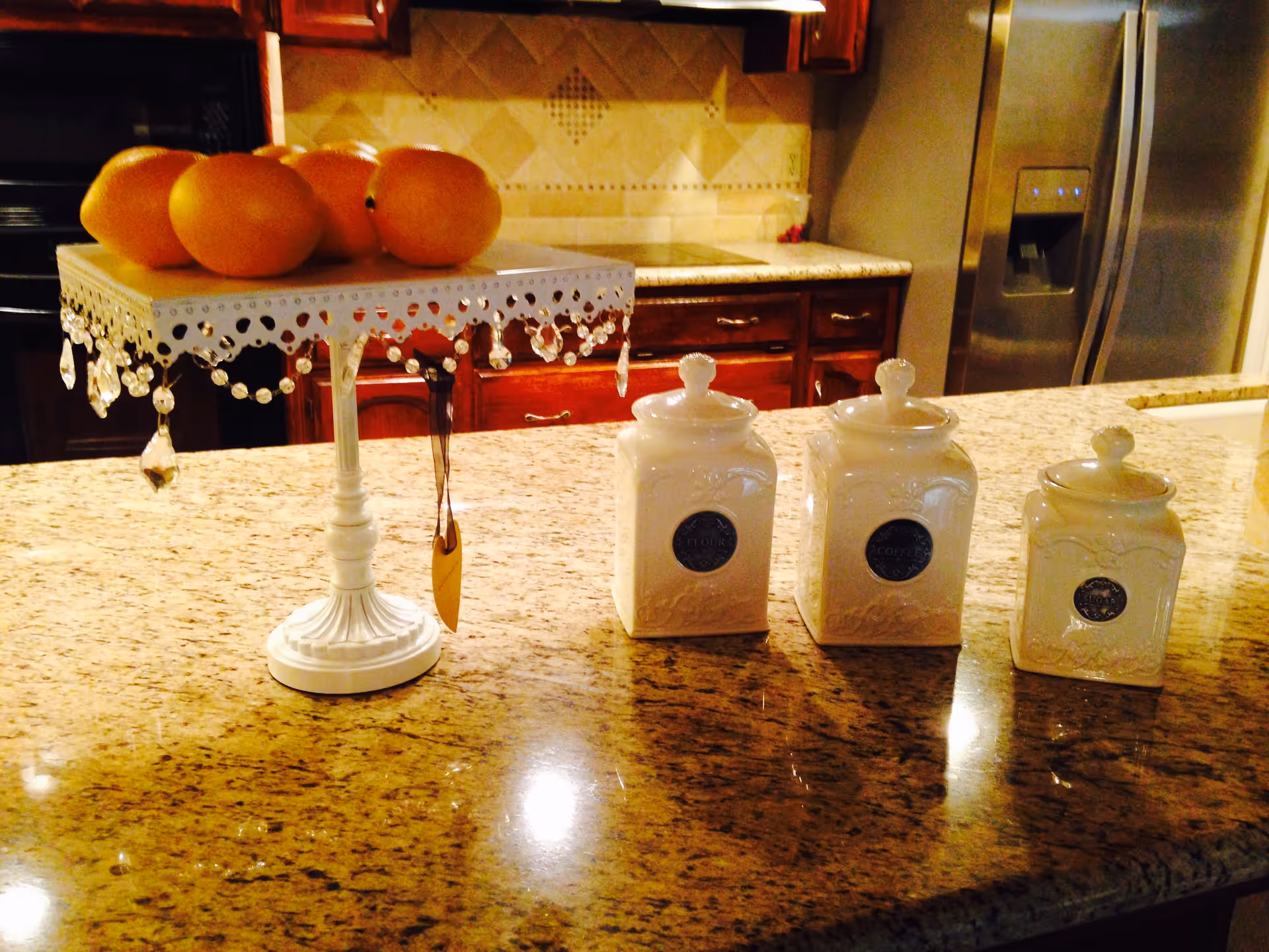 A kitchen countertop with a decorative white stand holding several oranges and three white ceramic canisters with lids arranged in a row. In the background, there are wooden cabinets, a tiled backsplash, and a stainless steel refrigerator.