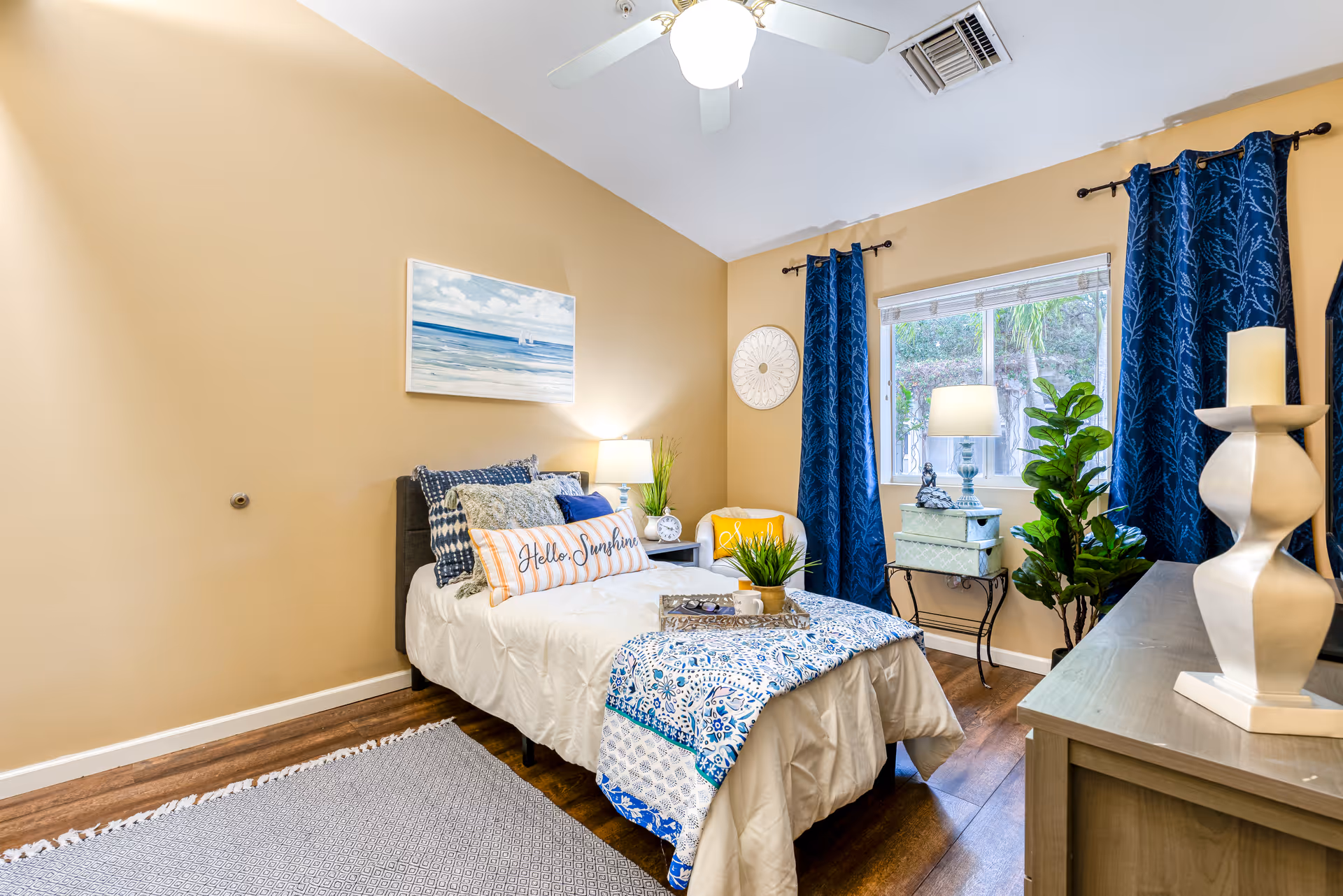 A cozy bedroom with a single bed adorned with multiple pillows including one that says 'Hello Sunshine'. The bed has a white comforter and a blue patterned throw blanket. There is a nightstand with a lamp and decorative items next to the bed. A window with blue curtains lets in natural light, and a small table with a lamp and decorative boxes is placed under the window. The room also features a ceiling fan, a yellow chair, a potted plant, and a dresser with a white candle holder on top. The walls are painted beige, and there is a beach-themed painting above the bed.