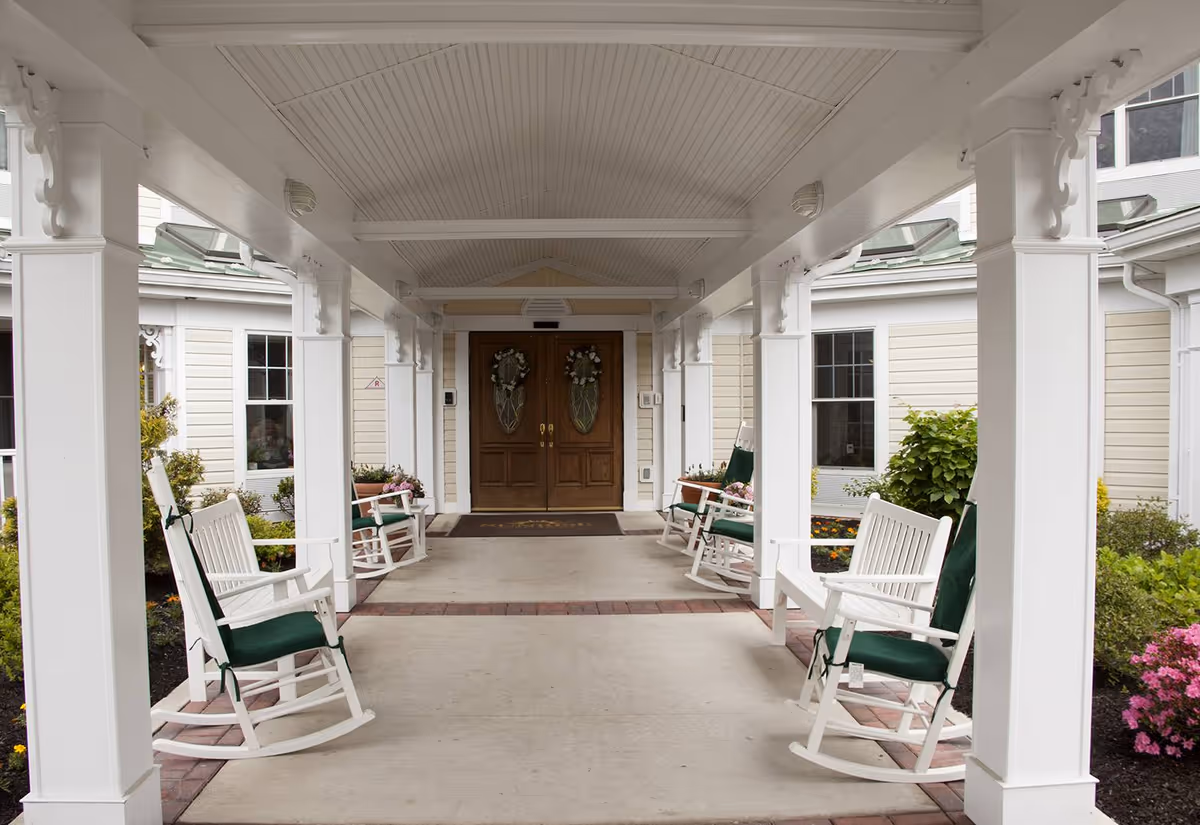 Covered entrance area of a senior living facility with white rocking chairs lined up on both sides, green cushions on the chairs, white pillars supporting the roof, and double wooden doors decorated with wreaths at the far end.
