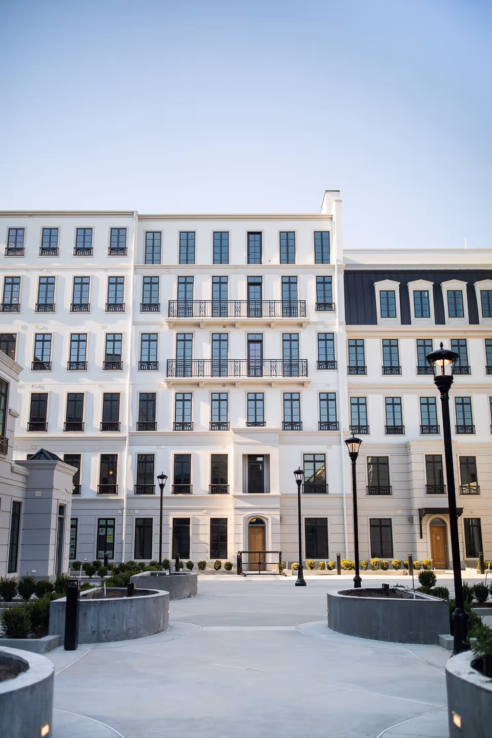 Exterior view of a multi-story white building with numerous windows and balconies, surrounded by a paved courtyard with modern street lamps and landscaped planters under a clear blue sky.