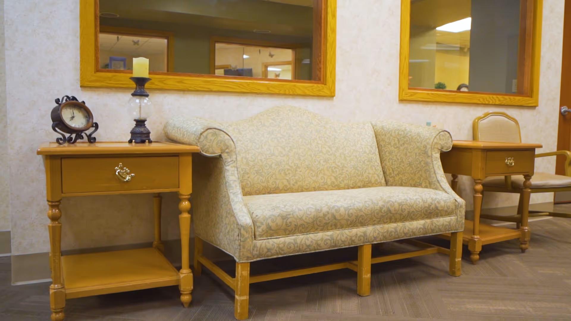A cozy seating area featuring a patterned beige loveseat flanked by two wooden side tables with drawers. One side table holds a decorative clock and a candle holder with a candle. Behind the loveseat are two large windows with wooden frames, reflecting the interior of the facility.