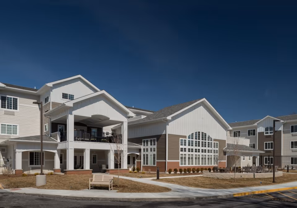 Exterior view of a multi-story senior living facility building with white and brown siding, large windows, and a covered balcony. There is a bench on the sidewalk and a small landscaped area with young trees and shrubs under a clear blue sky.