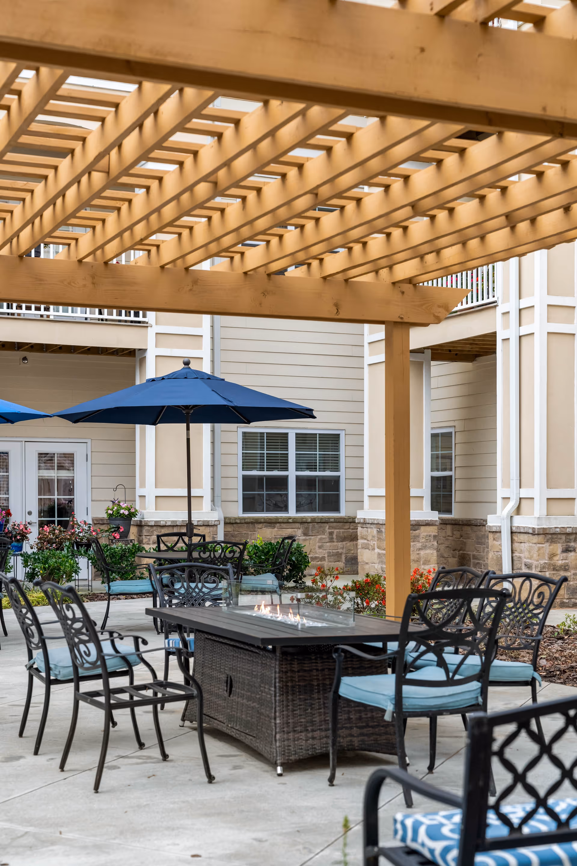 Outdoor seating area at a senior living facility with metal chairs featuring blue cushions arranged around a rectangular fire pit table under a wooden pergola. Blue patio umbrellas and a beige building with windows and stone accents are visible in the background.