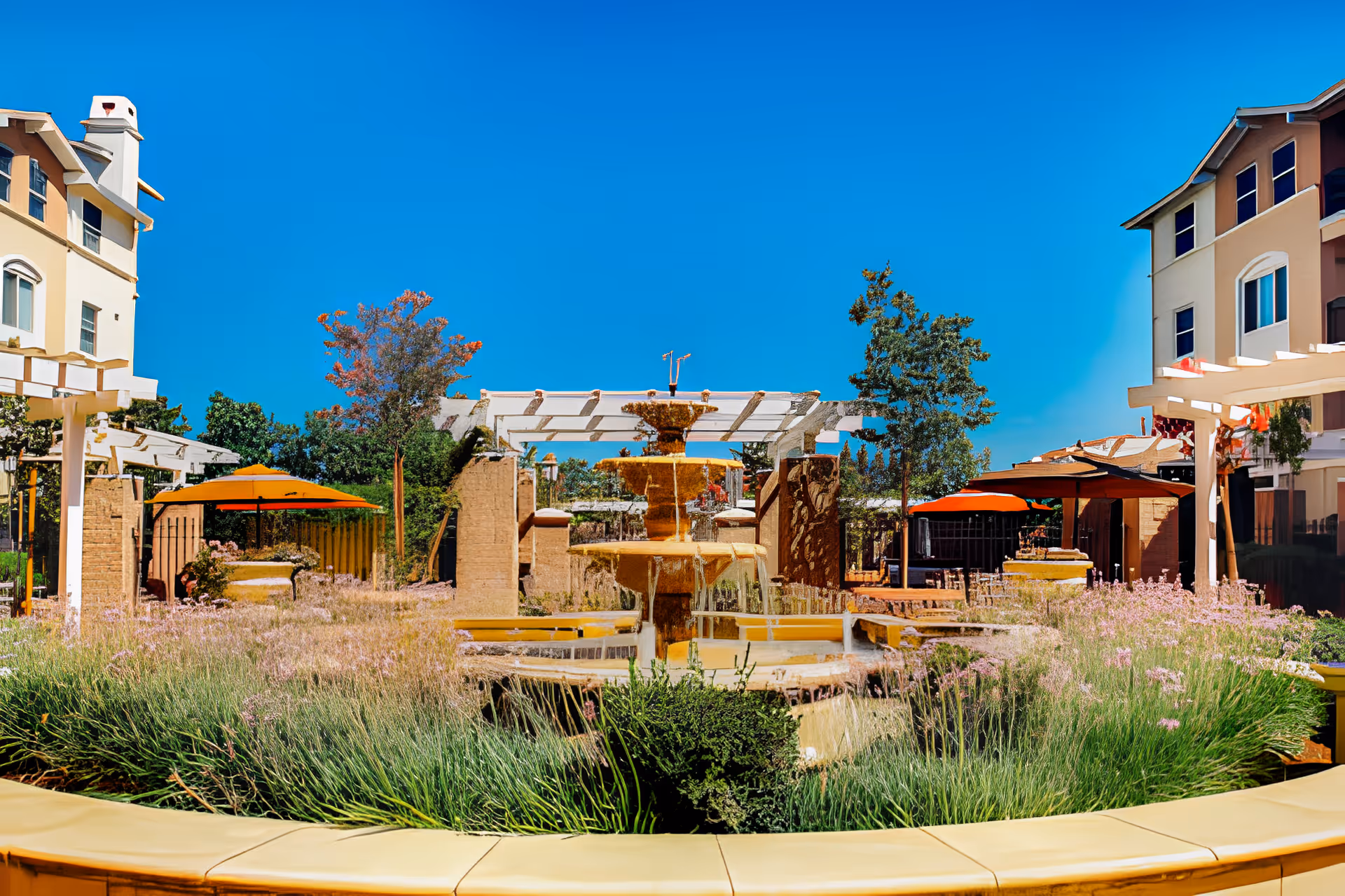 Outdoor courtyard area at Heritage Estate Senior Apartments featuring a central multi-tiered water fountain surrounded by lush greenery and flowering plants. There are pergolas and orange umbrellas providing shaded seating areas, with multi-story residential buildings in the background under a clear blue sky.