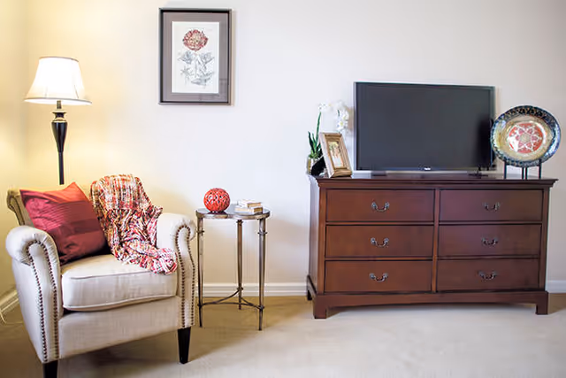 Cozy sitting area with an upholstered armchair, floor lamp, small side table, and a wooden dresser topped with a TV and decorative items.
