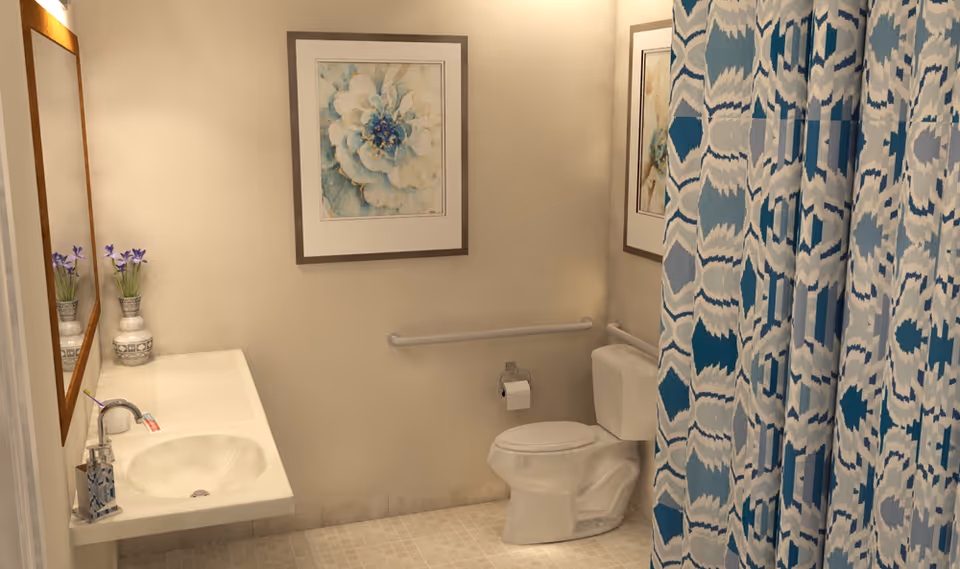 A clean and well-lit bathroom with a white sink and countertop on the left, a large mirror above the sink, a vase with purple flowers, a toilet with a grab bar on the wall behind it, a toilet paper holder, and a shower area with a blue and white patterned curtain. There are two framed floral artworks on the walls.