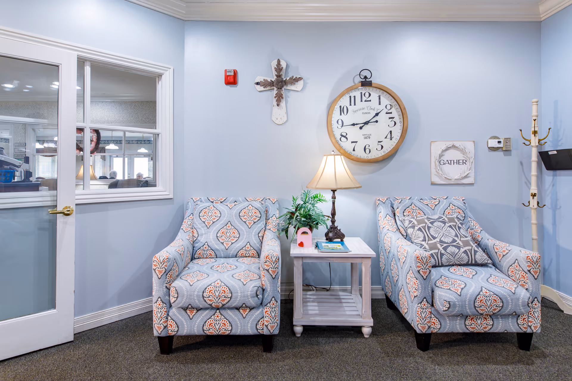 Two patterned armchairs flank a small white side table with a lamp and plant beneath a large wall clock and decorative art on a pale blue wall.