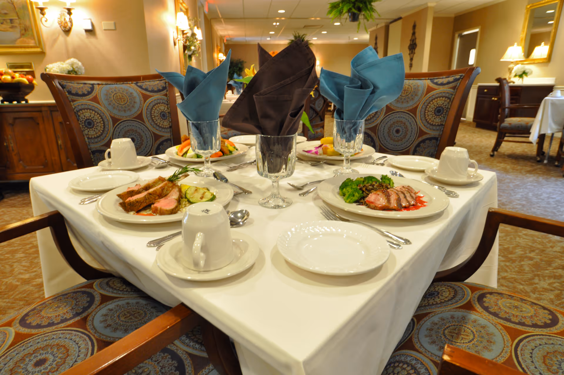 A formally set dining table with plated meals, folded napkins in glasses, cups, and patterned chairs in a dining room.