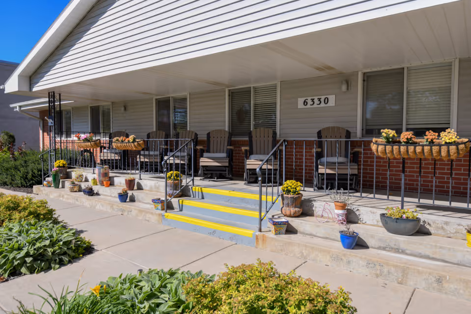 Front porch of a building with the number 6330 displayed on the wall. The porch has several cushioned chairs, black metal railings, and multiple flower pots with colorful flowers arranged along the steps and railing. The building exterior features beige siding and red brick.