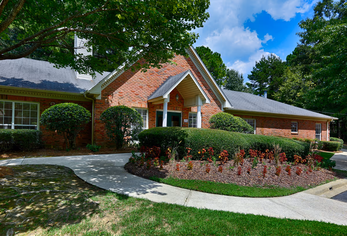 Brick single-story building entrance with a curved walkway, manicured shrubs and flowerbeds under leafy trees.