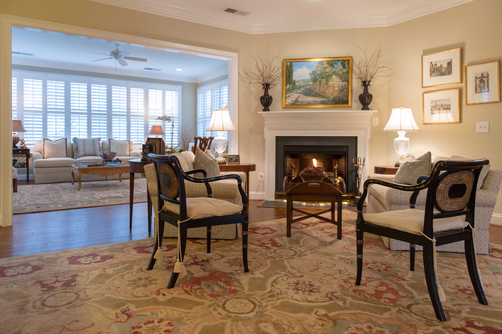 A cozy living room with two cushioned armchairs facing a fireplace with a fire burning. The fireplace mantel is decorated with two vases holding branches and a framed landscape painting. There are two table lamps on side tables next to a sofa. In the background, another sitting area is visible with a sofa, coffee table, and large windows with white shutters letting in natural light.