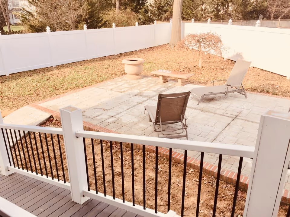 Outdoor patio area with a tiled floor, two lounge chairs, a small bench, a planter, and a small tree, all enclosed by a white fence with black vertical bars on the railing in the foreground.
