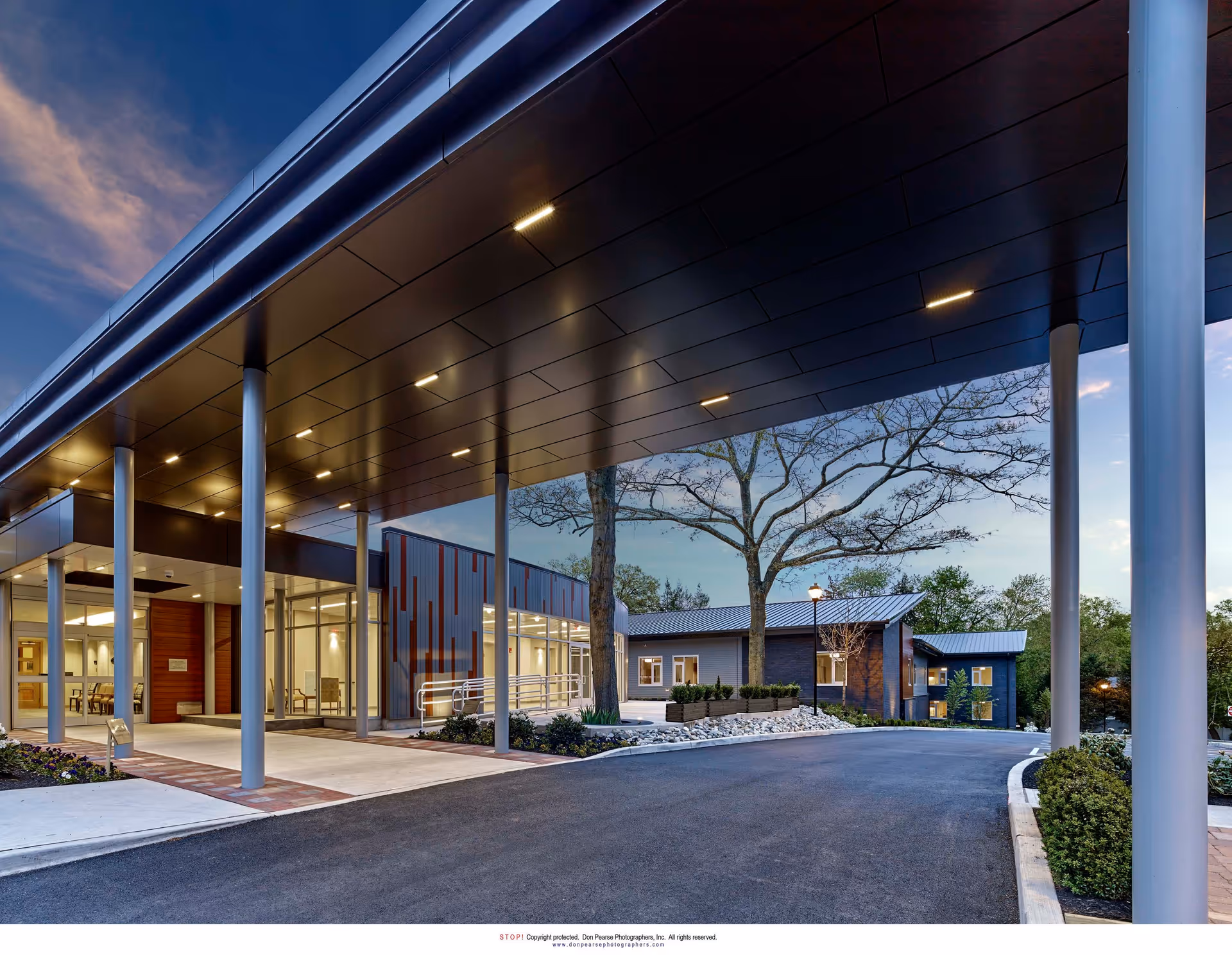 Exterior view of The Actors Fund Home building entrance at dusk, featuring a covered drop-off area with tall pillars, modern architecture, landscaped surroundings, and a clear sky with some clouds.
