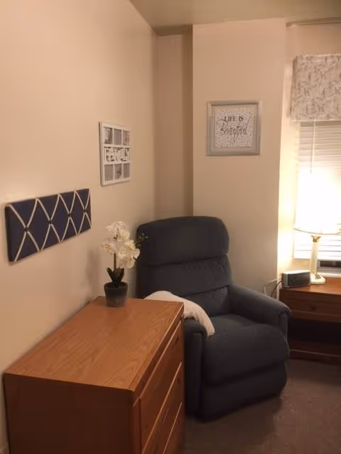 A cozy corner of a room featuring a dark gray recliner chair with a white pillow, a wooden dresser with a small potted white orchid on top, a framed inspirational quote on the wall that reads 'Life is Beautiful', a window with blinds and a floral valance, and a wooden nightstand with a lamp and a small black device.