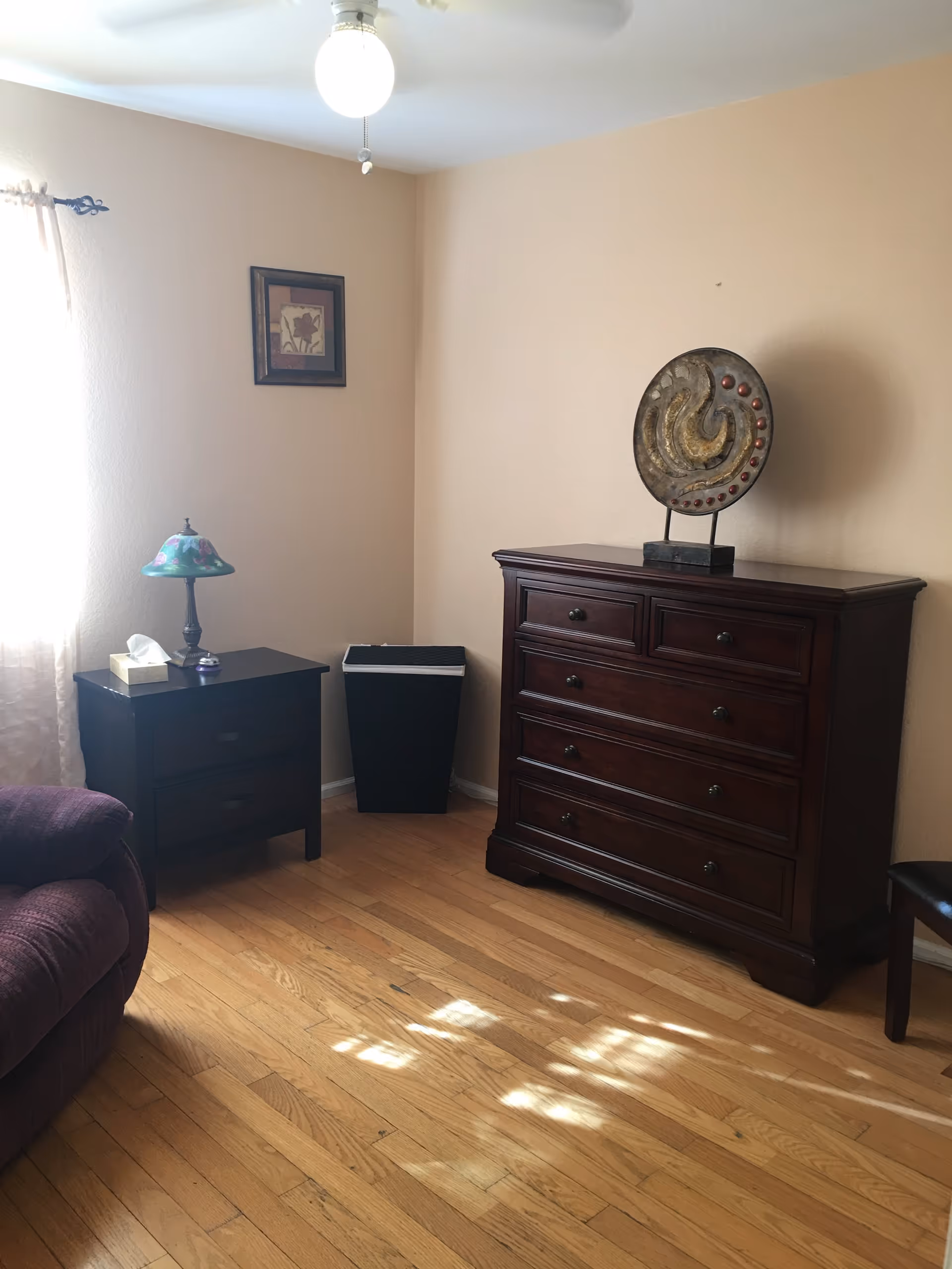A corner of a room with light wooden flooring and beige walls. There is a dark wooden dresser with five drawers and a decorative circular sculpture on top. To the left, there is a small dark wooden side table with two drawers, a floral lamp, and a tissue box. A purple upholstered chair is partially visible on the left side. A framed floral picture hangs on the wall above the side table. Sunlight streams in from a window with sheer curtains on the left.