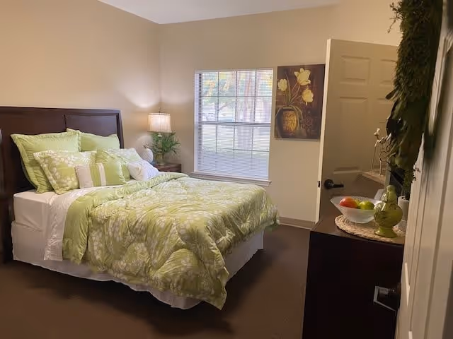 A cozy bedroom with a bed dressed in green and white floral bedding and multiple pillows. There is a wooden headboard, a nightstand with a lamp and a plant, a window with blinds, a floral painting on the wall, and a dresser with decorative items including a bowl of fruit and a green vase.