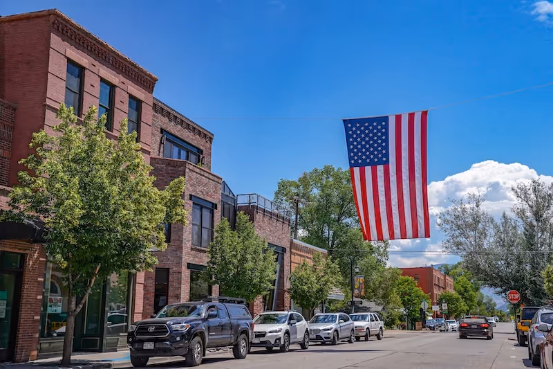 A brick building along a tree-lined street with parked cars and a large American flag hanging across the road.
