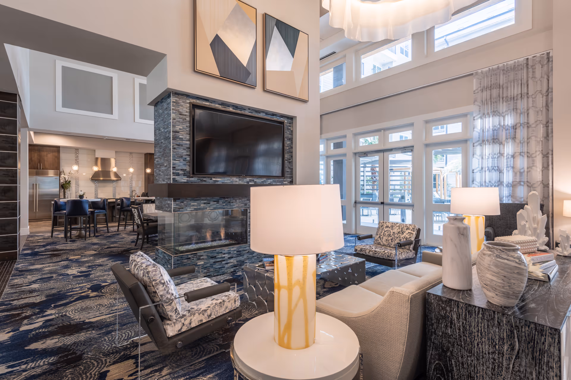 Bright, modern communal lounge with seating arranged around a central fireplace and wall-mounted TV beneath high windows.