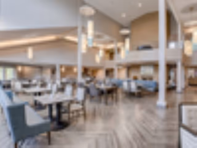 Spacious and well-lit dining area with multiple tables and chairs arranged neatly. The room features high ceilings with modern hanging light fixtures, large windows allowing natural light, and a mix of booth and chair seating. The flooring is wooden, and the overall decor is contemporary and inviting.