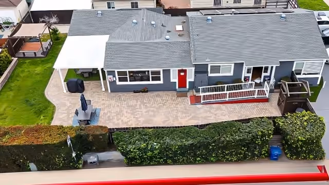 Aerial view of a single-story residential building with a gray roof and blue-gray exterior walls. The building has a red front door, a white covered patio area with outdoor furniture, and a wheelchair-accessible ramp. The surrounding area includes a paved patio, green hedges, and a small lawn.