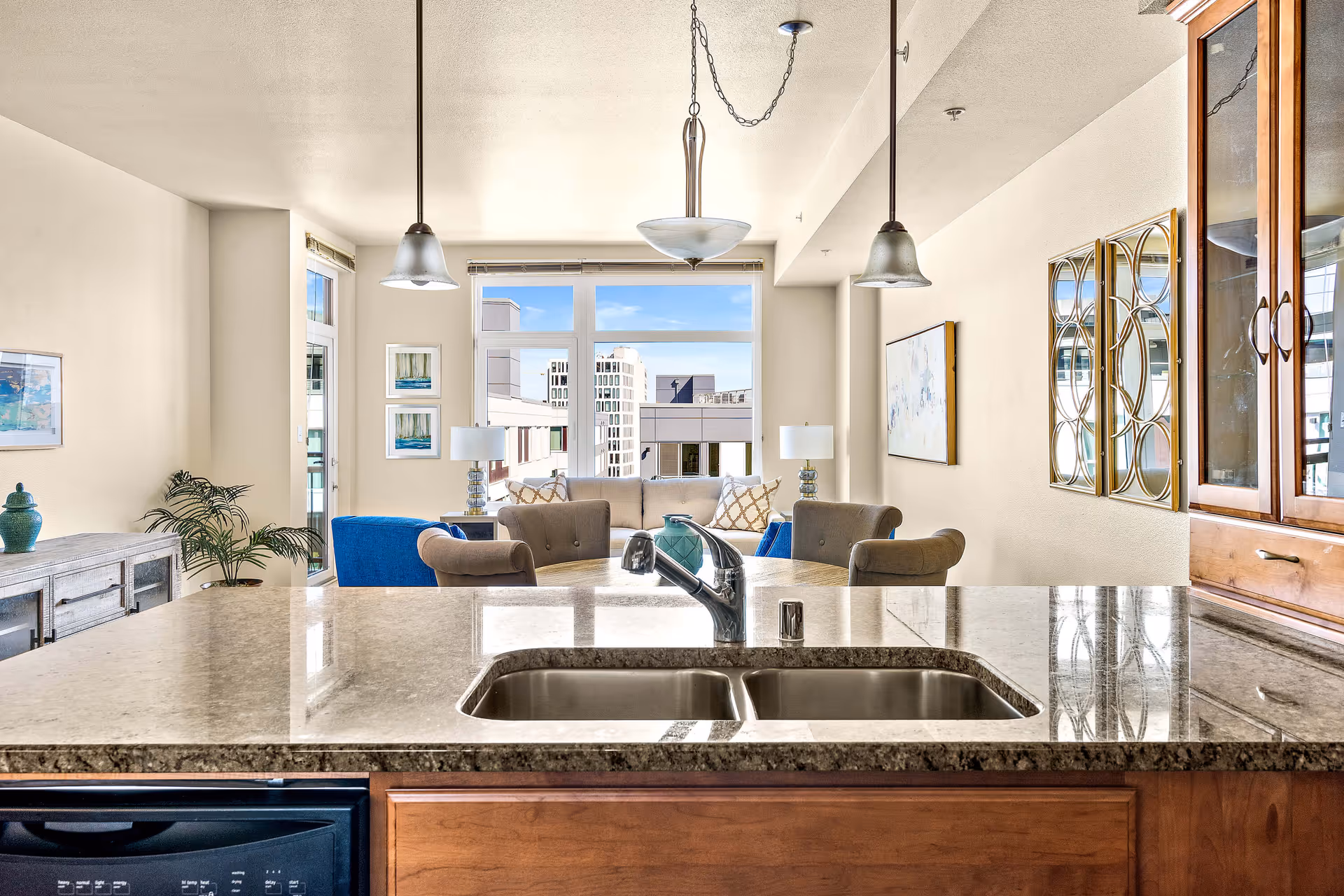 View from a kitchen island with a double sink overlooking a bright living room with sofas, pendant lights, and large windows.