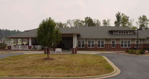Exterior view of a single-story senior living facility building with a covered entrance, brick and siding facade, a small tree in front, and a paved driveway.