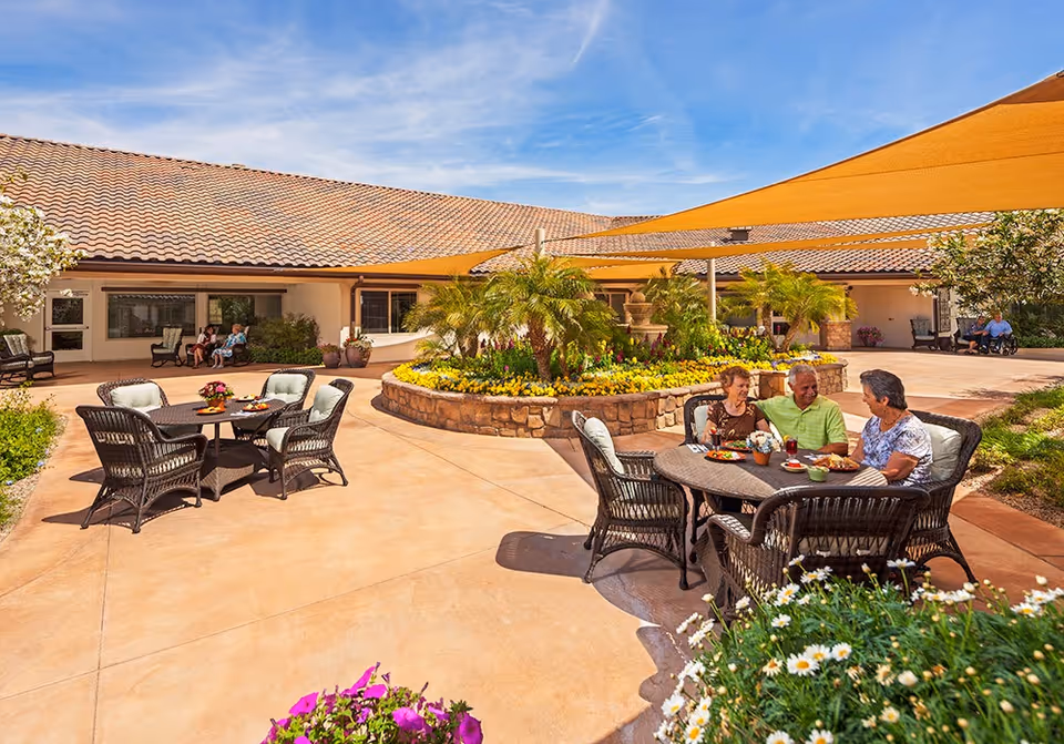 Outdoor courtyard area at Amber Creek Inn Memory Care Community with patio tables and chairs. Three elderly people are seated at one table enjoying a meal and conversation. The courtyard features a central raised flower bed with palm plants and colorful flowers, shaded by orange triangular sun sails. Additional seating and greenery surround the space under a clear blue sky.