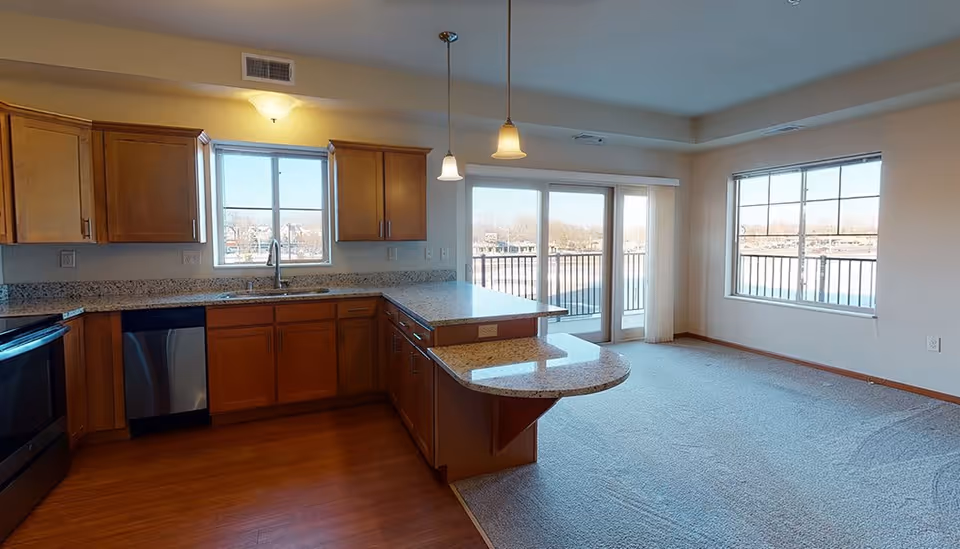 Interior view of a kitchen and adjacent living area in a senior living facility. The kitchen features wooden cabinets, granite countertops, a stainless steel dishwasher, and a stove. There is a window above the sink and two pendant lights hanging over the countertop. The living area has carpeted flooring and large windows with a sliding glass door that opens to a balcony with a view of a body of water and trees outside.