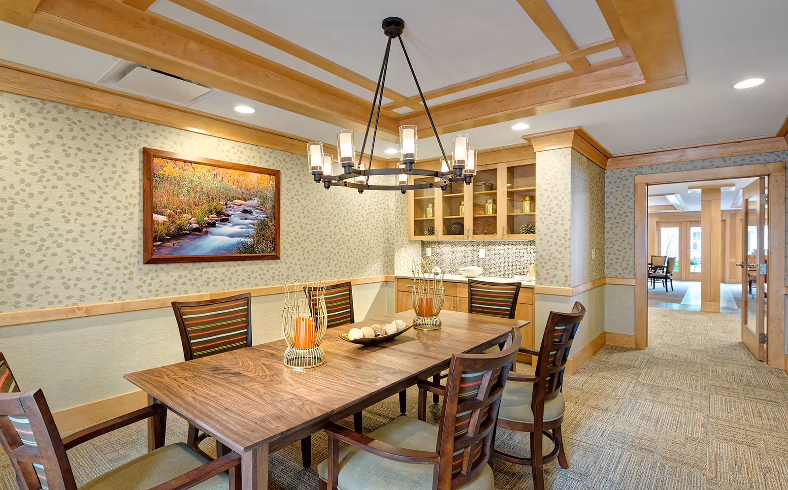 A well-lit dining room with a wooden table surrounded by six chairs with striped cushions. The room features a decorative chandelier hanging from a coffered ceiling, a framed landscape photograph of a stream on the wall, and built-in wooden cabinets with glass doors displaying decorative items. The floor is carpeted, and there is an open doorway leading to another room with more seating visible.