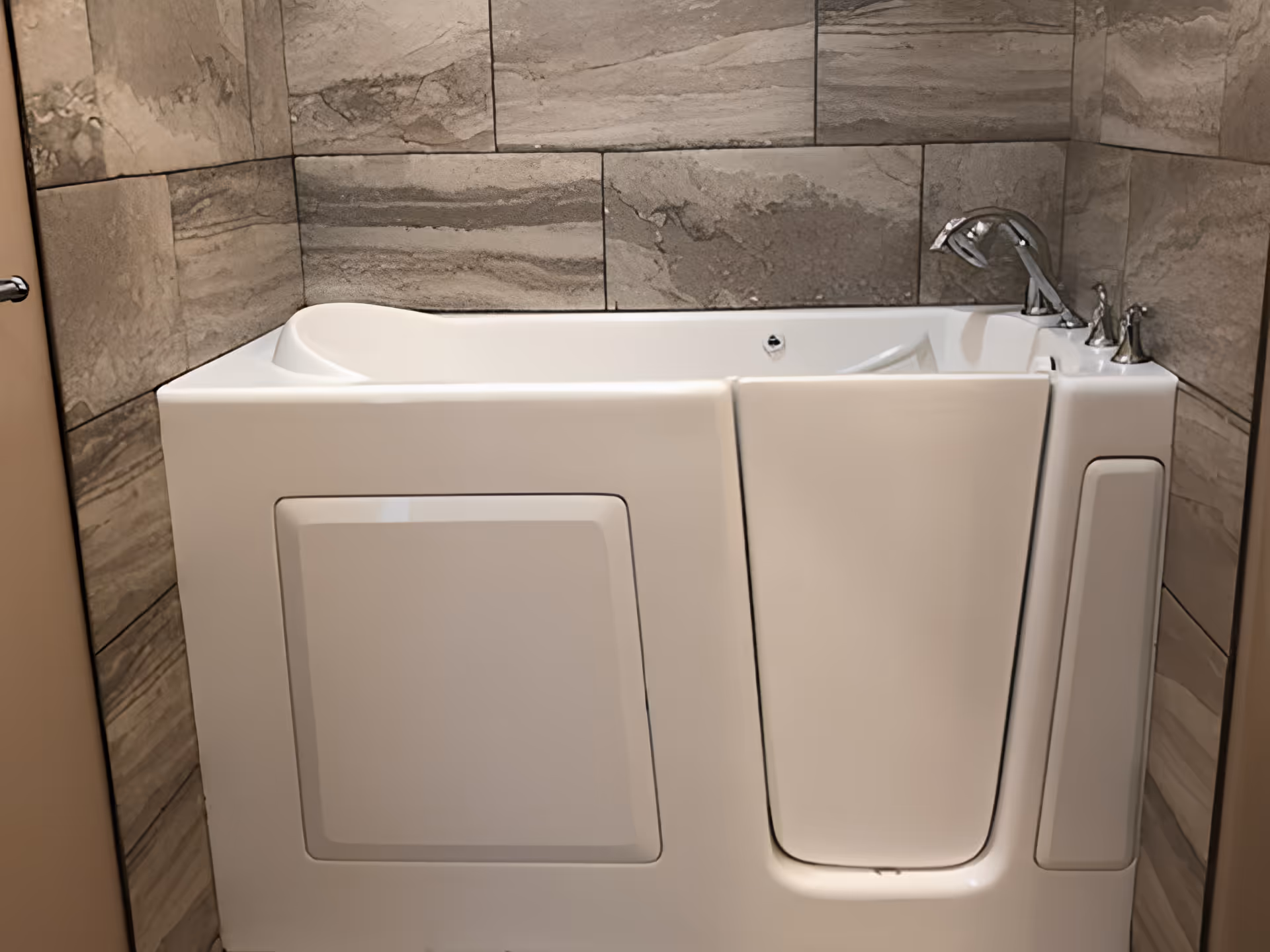 A white walk-in bathtub with a door, chrome faucet, and handles, surrounded by beige and gray stone tiles on the walls.