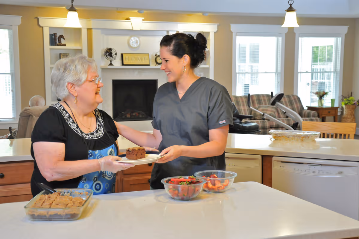 An elderly woman and a caregiver smile as they exchange a plate with cake in a bright communal kitchen area.