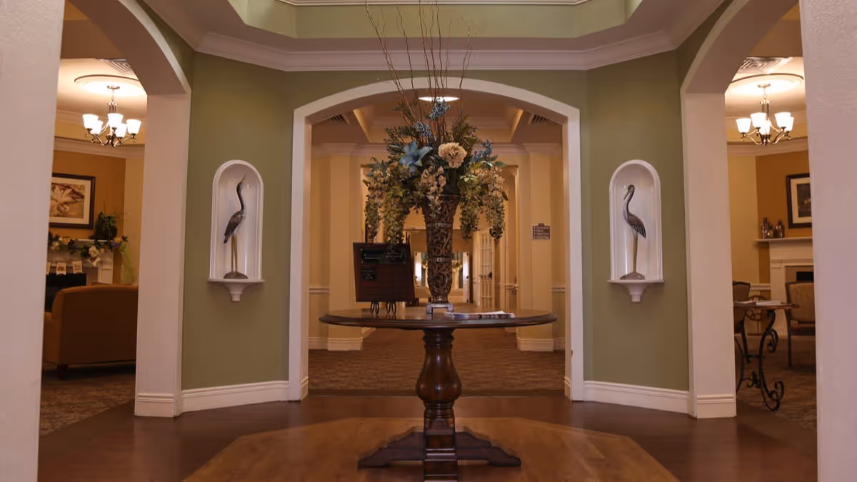 Interior view of a senior living facility lobby area with a round wooden table in the center holding a large floral arrangement. The walls are painted green with white trim, and two decorative niches each contain a bird sculpture. In the background, there are seating areas with chairs, tables, and framed artwork on the walls.