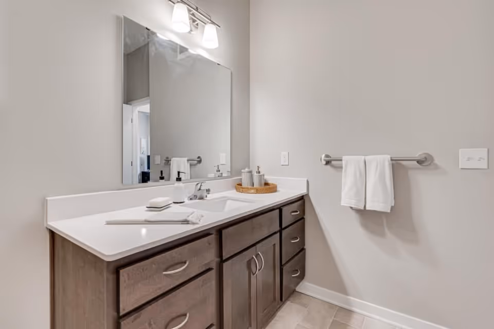 A bathroom vanity with a large rectangular mirror above it, a white countertop with a built-in sink, soap dispenser, soap bar, and decorative containers on a tray. There is a towel rack on the wall holding two white towels. The cabinetry is wooden with multiple drawers and cabinets.