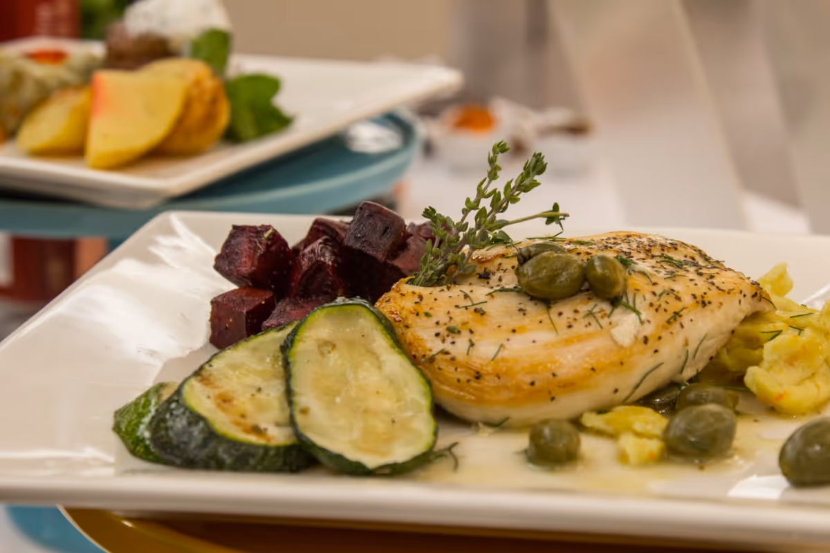 A plated meal featuring a piece of cooked chicken breast garnished with capers and herbs, accompanied by slices of cooked zucchini and diced beets on a white rectangular plate.