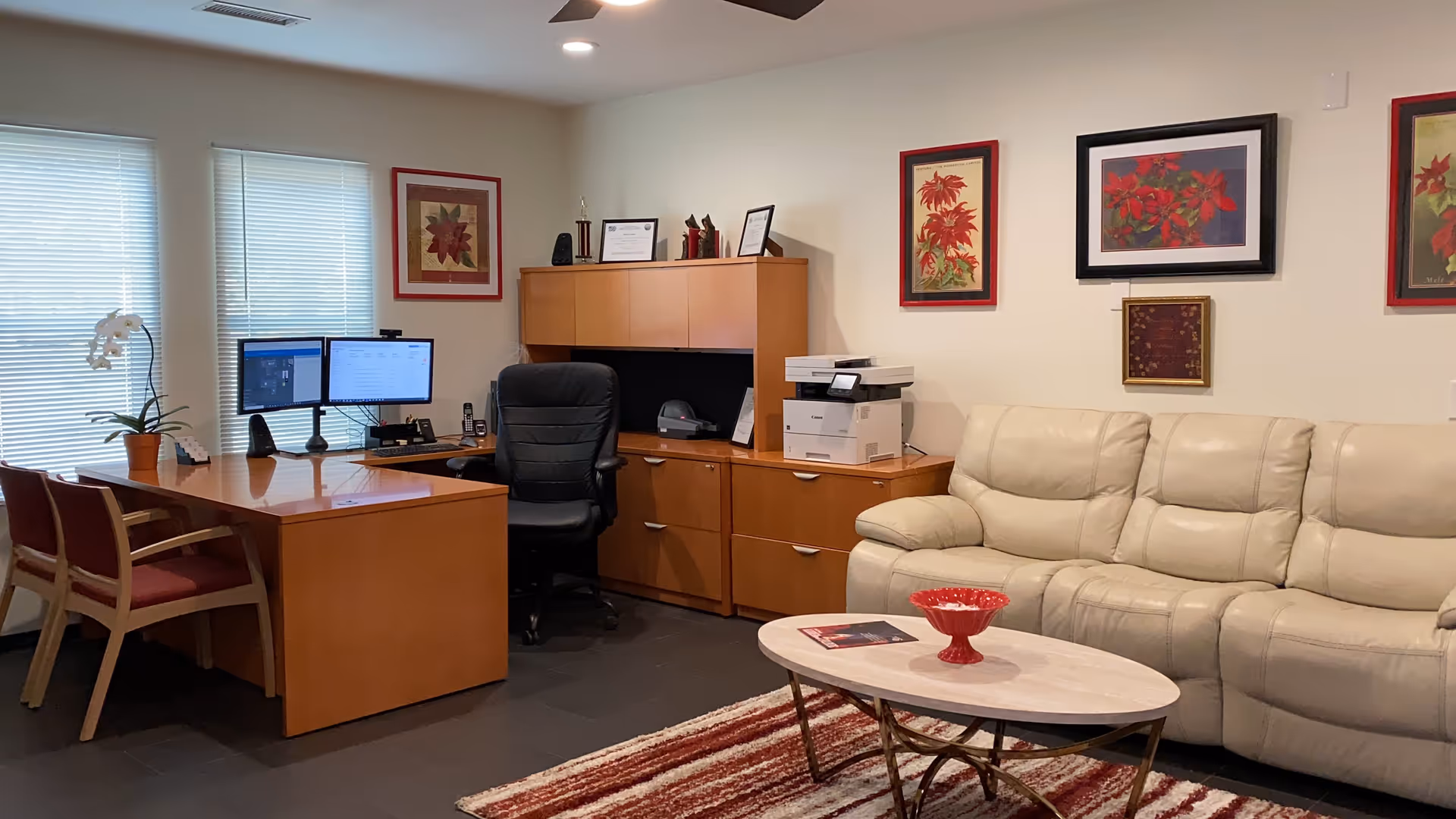 An office area with a wooden desk, two computer monitors, a black office chair, and a wooden cabinet with a printer on top. There is a beige leather couch and a small oval coffee table with a red decorative bowl. The walls have framed floral artwork, and two windows with blinds are visible.