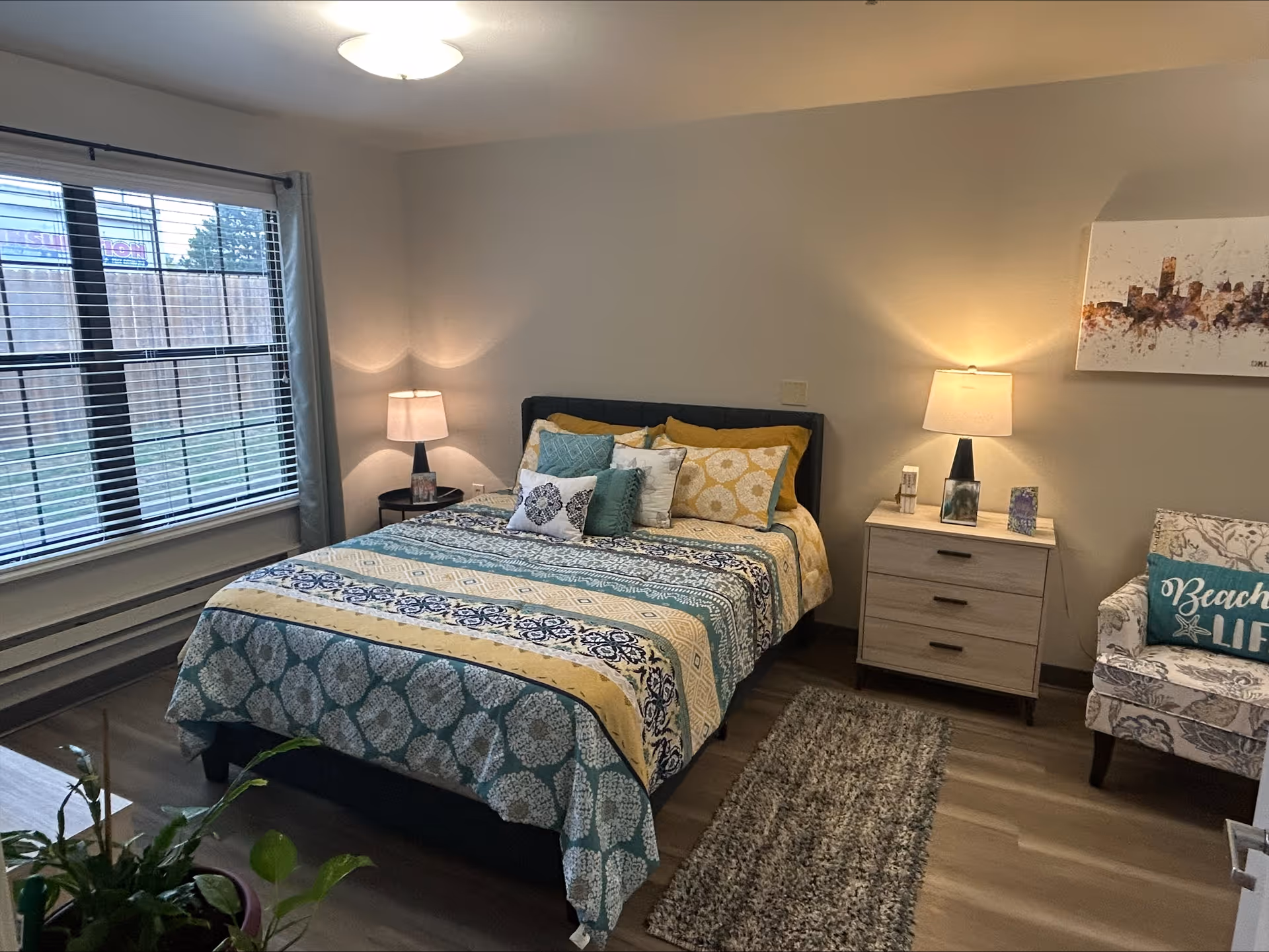 Decorated bedroom with a double bed dressed in patterned bedding, two nightstands with lamps, an armchair, a window with blinds and a small rug.