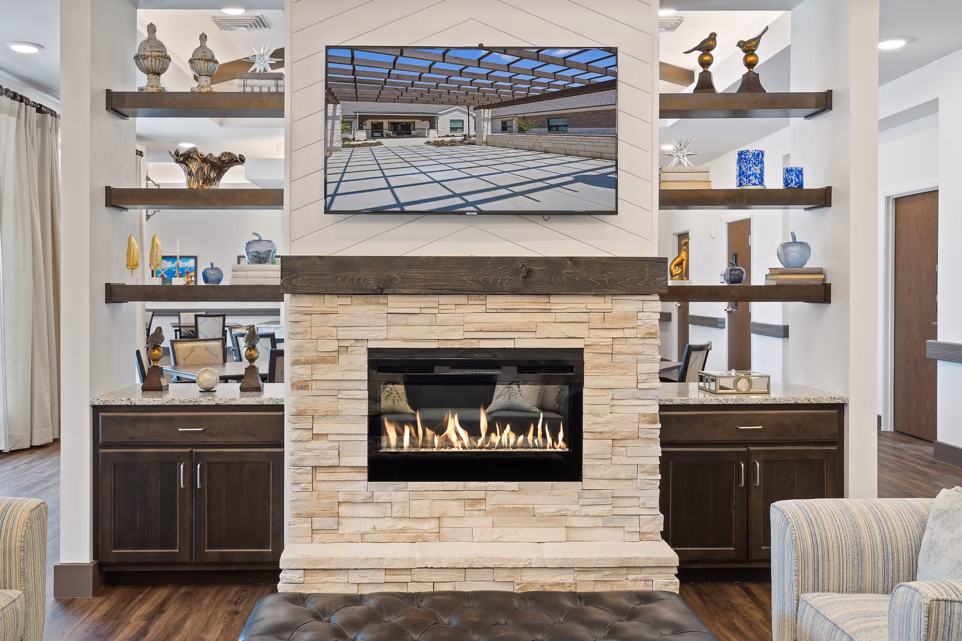 A cozy senior living common area featuring a modern stone fireplace with a wooden mantel and a flat-screen TV mounted above it. On either side of the fireplace are open shelves decorated with various vases, sculptures, and books. The room has wooden flooring and comfortable seating, creating a warm and inviting atmosphere.