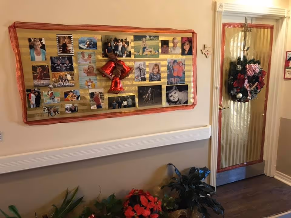 Interior hallway decorated for the holidays with a bulletin board displaying various family photos and a red bell decoration in the center. A door to the right is adorned with a Christmas wreath featuring red and silver ornaments and a striped bow. Several potted plants are placed below the bulletin board along the wall.