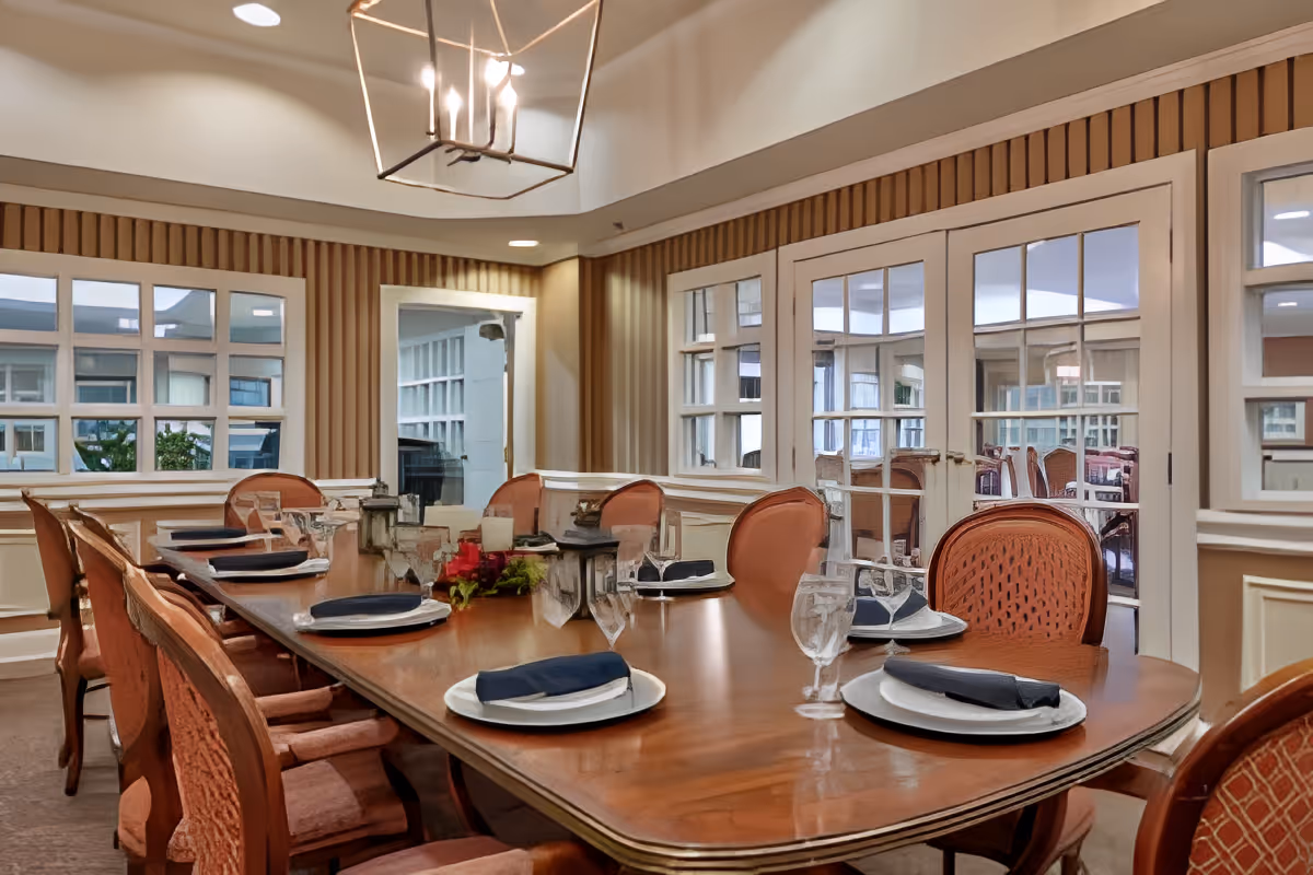 Formal dining room with a long wooden table set with plates, napkins, and glassware surrounded by upholstered chairs and windows with French doors.